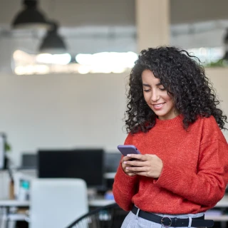 Smiling young professional latin business woman, happy lady corporate leader holding cellular phone working standing in modern office using mobile apps